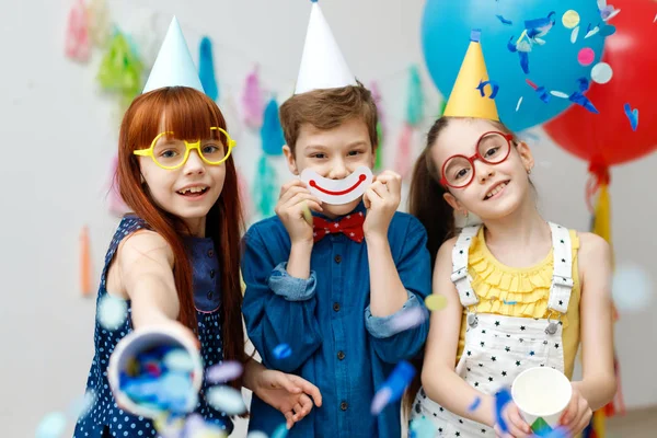 Tres niños amigables en gorras de cono festivo y grandes gafas, de pie en la sala decorativa con globos, divertirse juntos como celebrar el cumpleaños mirada con expresiones felices en la cámara, disfrutar de los juegos —  Fotos de Stock