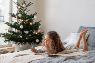 Uzun kıvırcık saçlı huzurlu küçük kadın çocuğun yatay atış dikkatle kağıdın görünüyor, farklı resimler renkler, dekore edilmiş Noel ağacı, tatil geliyor expectes yakınındaki yatakta yatıyor.
