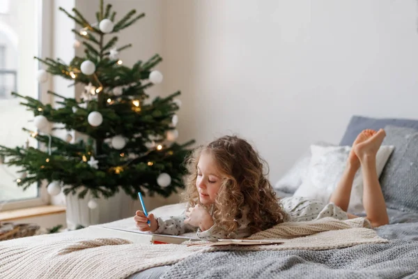 Horizontal disparo de descanso pequeño niño femenino con el pelo largo rizado mira con atención en la hoja de papel, colores diferentes imágenes, se encuentra en la cama cerca del árbol de Navidad decorado, las expectativas de las próximas vacaciones . — Foto de Stock