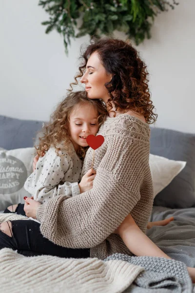 Tiro vertical de cariñosa joven madre y su hija se abrazan y expresan amor, se sientan en una cama cómoda, cierran los ojos con placer, se relajan y se complacen. Concepto de paternidad . — Foto de Stock