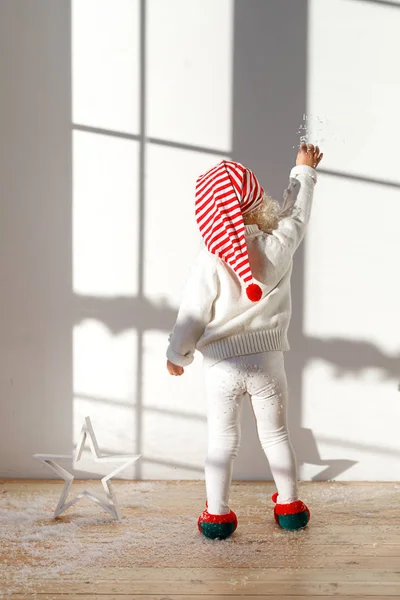 Back view of adorable small kid elf wears white costume, santa s hat and elf s shoes, plays with artificial snow, stands on wooden floor in spacious room, likes playig. Children, celebration concept