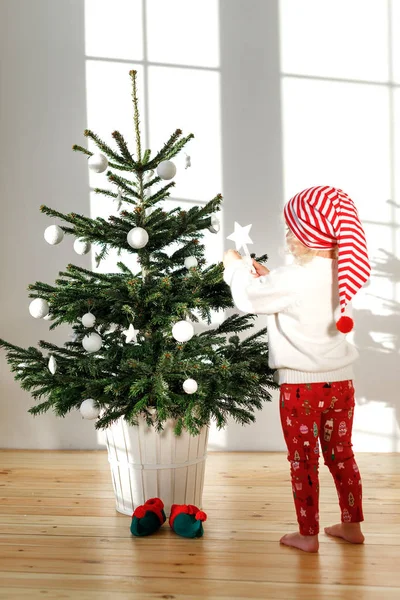 Retrato vertical de una niña rubia pequeña con los pies desnudos en el suelo de madera, decora el árbol de Año Nuevo, usa sombrero de Santa Claus, le gusta la preparación para las vacaciones. Concepto infantil y navideño . — Foto de Stock