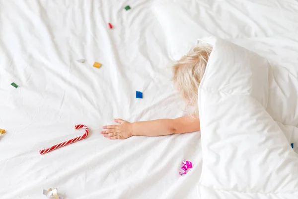 Chica bastante adorable se encuentra debajo de una manta blanca cálida, tiene sueños, estratifica la mano a dulces caramelos, disfruta del descanso en la cama. Precioso niño pequeño duerme en el dormitorio, disfruta del descanso. Los niños y el ocio . — Foto de Stock