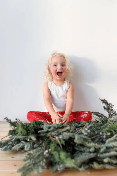 Alegre niña rubia alegre se sienta piernas cruzadas y guirnalda de Navidad en primer plano, aislado sobre fondo rosa. Adorable niño sonriente pasa tiempo en casa con los padres de buen humor — Foto de Stock