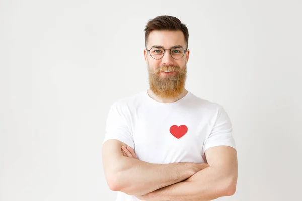 Portrait of handsome bearded young man wears round spectacles and white casual t shirt with red heart, keeps hands crossed, looks confidently into camera, isolated over white studio backgorund — Stock Photo, Image