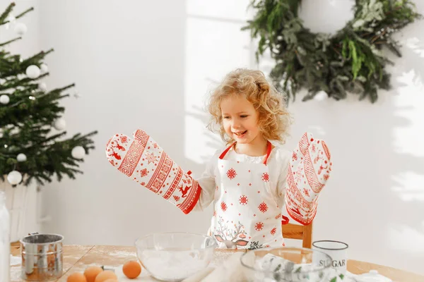 Trabajador niño pequeño cocinero usa guantes de cocina grandes, prepara deliciosa pastelería en lugar de la madre, confiado en el éxito, ayuda a los padres sobre la casa, se encuentra en la habitación espaciosa decorada con abeto . — Foto de Stock