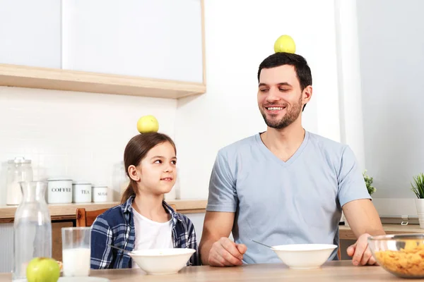Funny young dad and daughter sit next to each other, eat delicious cornflakes, keep apples on head, demonstrate that they eat only healthy food, have good relationships and understanding.