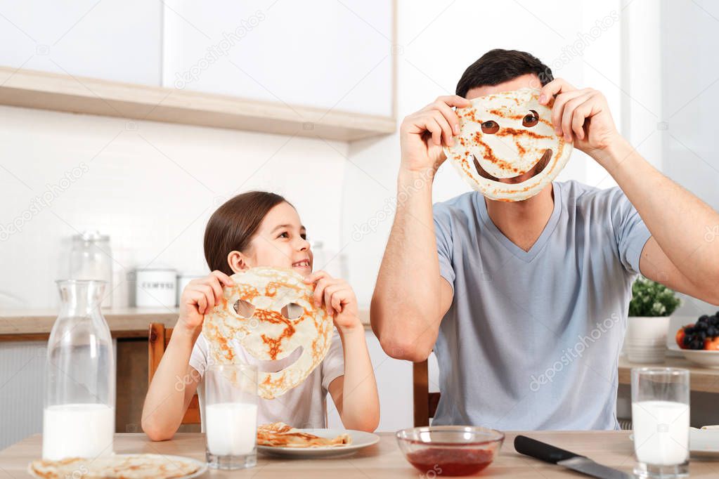Glad positive young father foolishes with her small daughter at kitchen, make faces from pancakes, have tasty breakfast, pose at camera, drink fresh milk, eat flannel cakes with sweet jam.