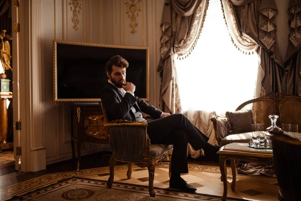 Elegant prosperous businessman in formal black suit, sits on chair in royal room, feels relaxed, has serious thoughtful expression as thinks about future plans, has thick beard and stylish hairdo.