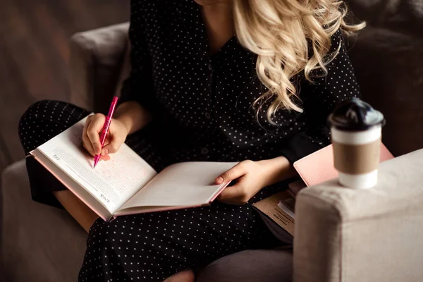 Cropped shot of unrecognizable blonde female writes down information in notebook or diary, prepares for examination, drinks hot tasty beverage, spends time at home. Woman notes something in notepad. — Stock Photo, Image