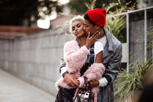 Amor mulato pareja abrazarse mientras tener caminar al aire libre, beso. Hombre elegante de piel oscura en sombrero rojo de moda abraza a su novia con apariencia específica. Gente, ética, concepto de relación — Foto de Stock