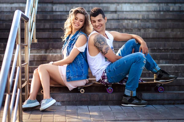 Cheerful girlfriend and boyfriend sit together backs on stairs, have rest after going skateboarding in open air, satisfied with summer weather, dressed in fashionable clothing. Active lifestyle. — Stock Photo, Image