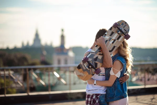 Casal encantador de skatistas ativos abraçar e beijar apaixonadamente, esconder-se atrás de skate, ficar contra a maravilhosa vista panorâmica da cidade com muitos lugares de interesse no fundo . Imagens Royalty-Free