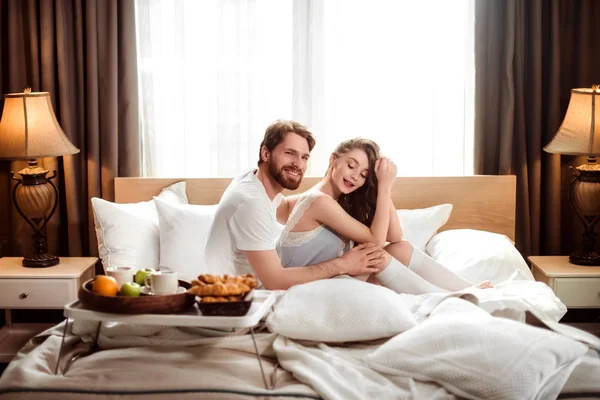 Улыбающийся бородатый мужчина счастлив проводить свободное время со своей возлюбленной, сидеть вместе в удобной постели в гостиничном номере, завтракать. Роскошные апартаменты. Понятие любви и отношений . — стоковое фото