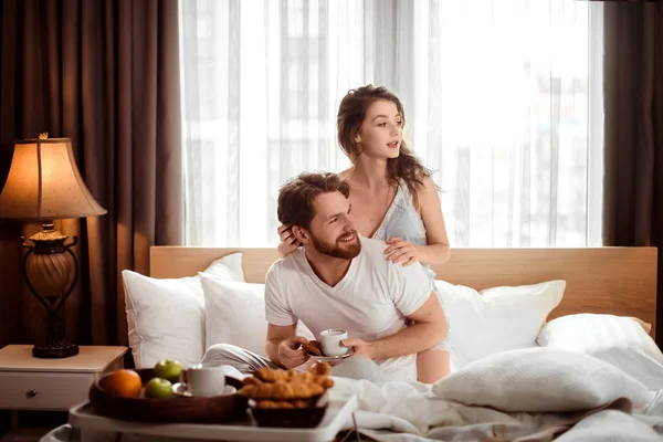 Satisfeito casal familiar tomar café da manhã na cama, desfrutar de deliciosos croissants, café e frutas, olhar positivamente afastado, senta-se contra aconchegante interior doméstico no quarto. Hora de dormir e conceito de relacionamento Fotografia De Stock