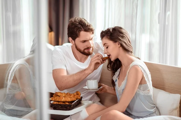 Conceito de amor e cuidado. Carinhoso jovem bonito barbudo macho alimenta sua namorada bonito com croissant, sentar juntos na cama confortável. Mulher bonita com bebida gosta de cuidar de seu namorado Fotografia De Stock