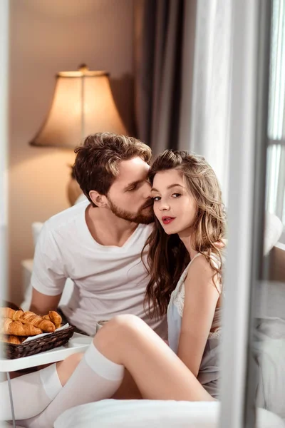 Tiro vertical de agradável olhando fêmea e seu bonito namorado barbudo que a beija na bochecha, desfrutar juntos deliciosos croissants, sentar no quarto de hotel de luxo, recriar no exterior durante as férias Fotografia De Stock