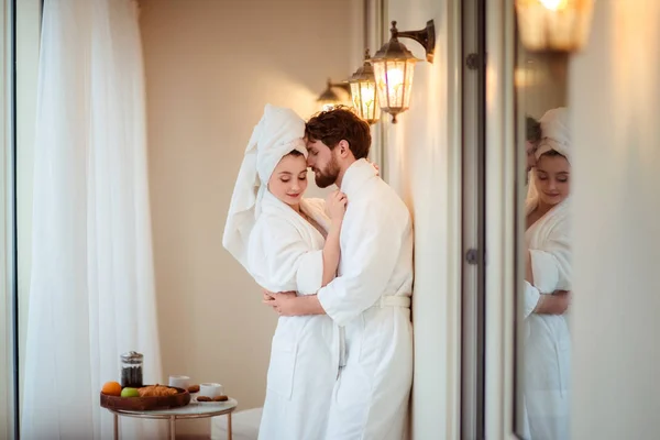 Homem barbudo e sua esposa usam roupões de banho brancos e toalha na cabeça, se abraçam, se sentem relaxados depois de tomar banho no hotel, indo dormir. Mulher adorável e seu marido posar após o chuveiro Fotografias De Stock Royalty-Free