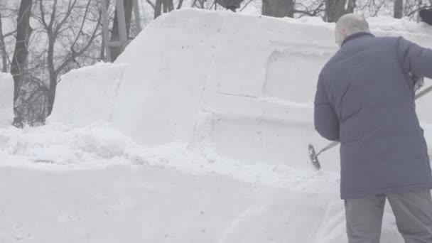 Mann schaufelt Schnee. Skulpturen aus Schnee herstellen. Schneeskulptur, Blöcke, die darauf warten, geformt zu werden. Schneeskulpturen von Tieren bei künstlicher Beleuchtung. Mann mit Schneeschaufel reinigt Gehwege in — Stockvideo