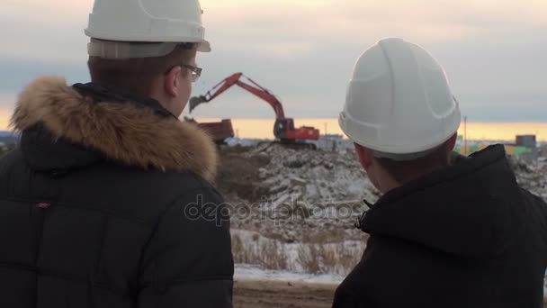 Baksidan på arkitekter inspektera byggarbetsplatsen. Två ingenjörer i hårda hattar på konstruktion webbplats grävmaskin i bakgrunden. Tekniker undersöker handlingar på byggarbetsplatsen mot grävmaskin. — Stockvideo
