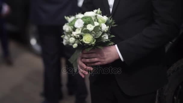 Un hombre de negocios guapo con flores. Novio en un traje con un ramo de flores. Boutonniere de boda. Elegante hombre vestido con un traje negro moderno y camisa blanca sosteniendo en la mano flores de boda — Vídeos de Stock