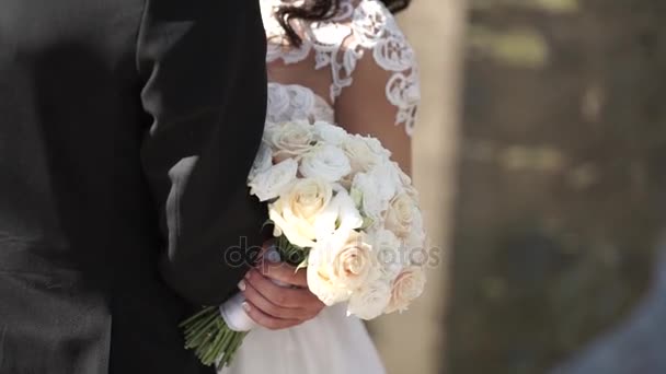 Elegante bruid en bruidegom poseren samen buiten op een trouwdag. Bruid een witte roos boeket te houden terwijl de volgende permanent aan de bruidegom. bruid in een jurk permanent en houden van een bruiloft boeket — Stockvideo