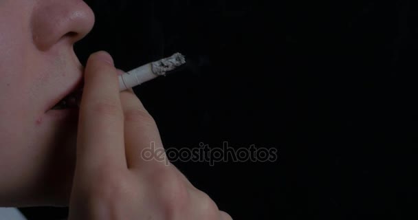 Человек курит сигарету на черном фоне. Закрыть портрет молодого человека, курящего сигарету. Курение. Молодой человек курит сигарету — стоковое видео