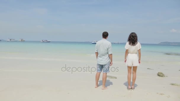 Pareja joven de pie sobre arena mojada por el mar y mirando a un cielo azul. Una hermosa joven sostiene a su guapo compañero mientras observan el atardecer juntos en una playa romántica. Vacaciones de verano — Vídeos de Stock