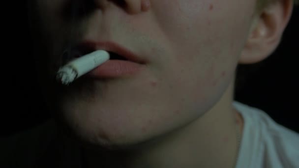Парень в белой футболке курит сигарету на черном фоне крупным планом. парень курит на чёрном фоне — стоковое видео