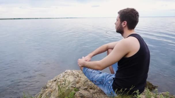 Young man sitting on a rock cliff above a rural lake. Country boy. Lonely young man — Stock Video