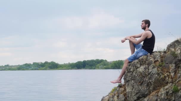 Mladý muž sedí na skalní vyvýšenině nad venkovských jezero. Vesnický chlapec. Osamělý mladý muž — Stock video