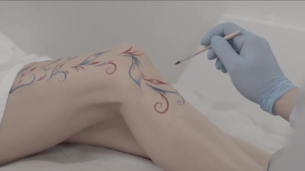 Närbild av en konstnär hand ritning tillfällig tatuering på någon ben. Unga kvinnliga kroppskonst. Målare gör en prydnad och dekoration på hennes ben — Stockvideo