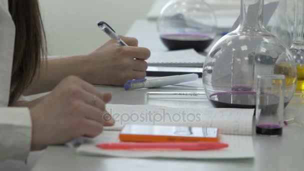 Los estudiantes que trabajan en el laboratorio de química toman notas en un cuaderno. Alumna usando el teléfono en la lección de ciencias. Jóvenes estudiantes de medicina escriben — Vídeo de stock