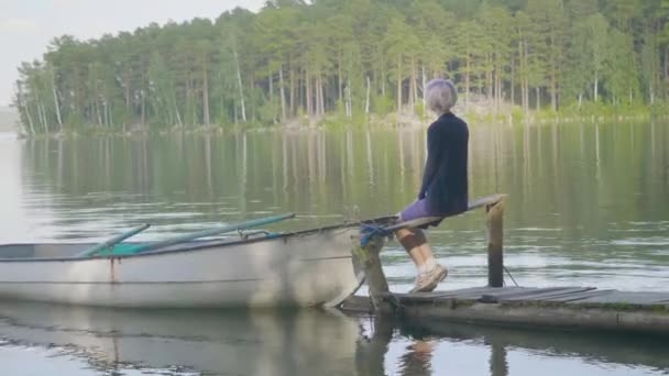 Vista laterale di una ragazza seduta sul molo di legno sulla riva del fiume. Vista laterale di sognare ragazza seduta sul molo di legno sulla riva del fiume. Bella donna con i capelli biondi — Video Stock