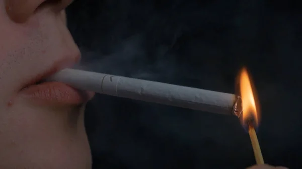 Человек курит сигарету на черном фоне. Закрыть портрет молодого человека, курящего сигарету. Курение. Молодой человек курит сигарету — стоковое фото