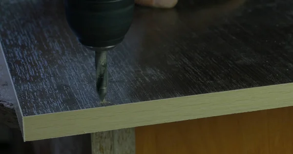 Плотник с помощью электрической дрели проделывает отверстия на доске на столе столярной мастерской. the drill makes a hole — стоковое фото