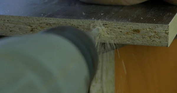 El hombre sostiene un taladro en sus manos y hace un agujero en una tabla de madera. Afeitaciones de madera volando alrededor. De cerca. — Foto de Stock