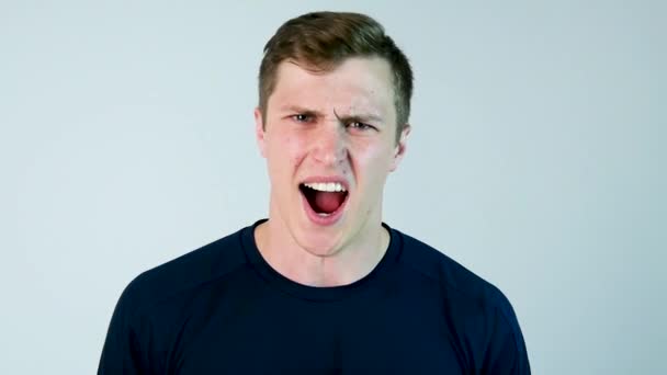 Een jonge man schreeuwt tegen de camera met zwart t-shirt en een grijze achtergrond. Slow motion — Stockvideo