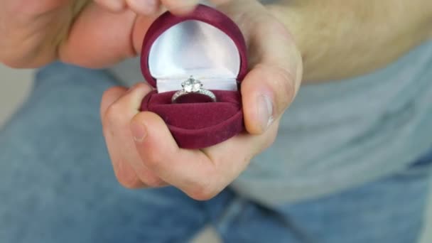 Primer plano del hombre con caja de regalo y anillo de compromiso sobre fondo de luces navideñas. Anillo de compromiso en las manos. Mano masculina abre caja roja con anillo de compromiso en el interior sobre fondo blanco — Vídeos de Stock