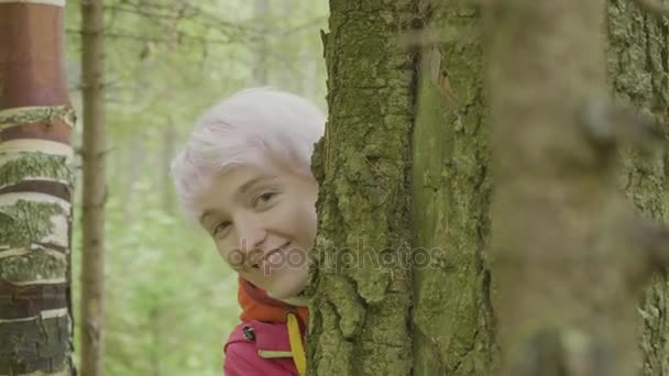 Jovem mulher sorridente atraente olhando atrás da árvore. Menina loira verificar e olha de trás de uma árvore Na floresta de outono — Vídeo de Stock