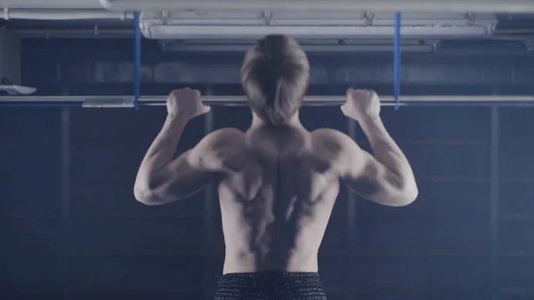 Back view of muscular man with naked torso doing pull ups exercise on horizontal bar. Fitness, gymnastics workout in gym. Man with naked torso is pulling on the bar back view