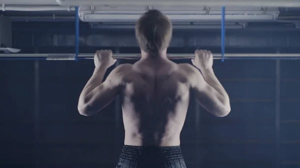 Back view of muscular man with naked torso doing pull ups exercise on horizontal bar. Fitness, gymnastics workout in gym. Man with naked torso is pulling on the bar back view