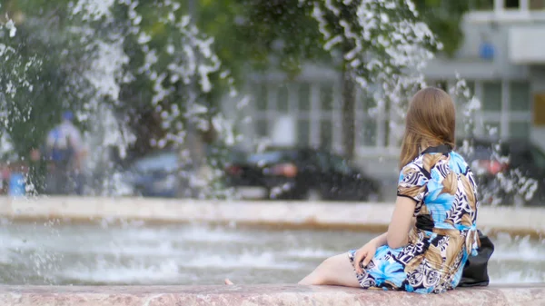 Kız oturan, arka planda akan bir çeşme. Kız çeşme tarafında oturan görünüm