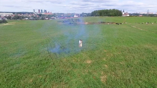 Aerial survey of couple walk on the field with colored smoke in hands. Flying over man and woman run through the field using colored smoke.Lovers running through the field with red and pink smoke