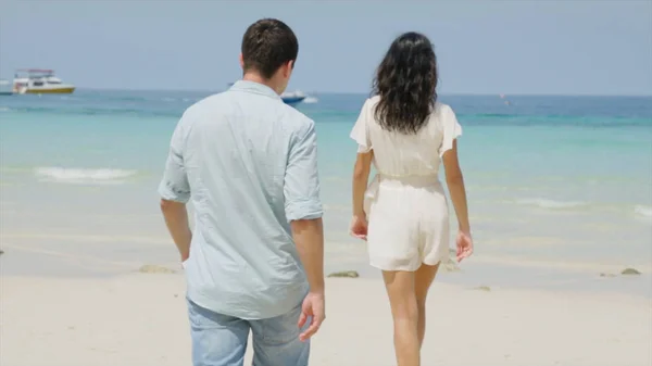 Couple mignon en vêtements blancs va plage de main. Deux adolescents qui courent et flirtent sur la plage près de l'eau. Femme marchant sur la plage vacances tenant la main du petit ami qui la suit — Photo