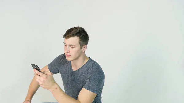 Nuori komea mies istuu puhelin kädessä valkoisella taustalla — kuvapankkivalokuva