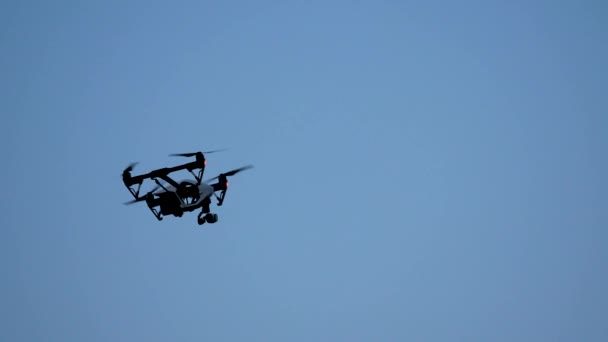 Черный квадрокоптер с камерой, летающей над голубым небом. Черный дрон в небе. Летающий квадровертолетный дрон в небе с установленной цифровой камерой для видео- и фотопроизводства — стоковое видео