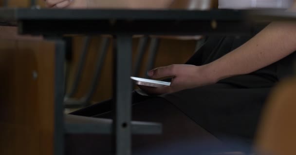 Mão de mulher usando um telefone inteligente sob uma mesa de madeira. Mulher usa a Internet no seu telefone durante o exame — Vídeo de Stock