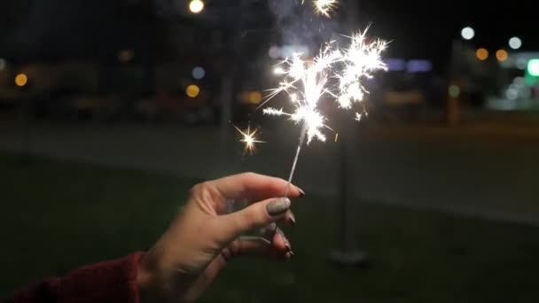 Lindo sparkler na mão mulher no fundo preto ou rua. Mulher segurando sparkler contra luzes coloridas desfocadas, de perto. Mãos femininas segurando sparkler no fundo escuro — Vídeo de Stock