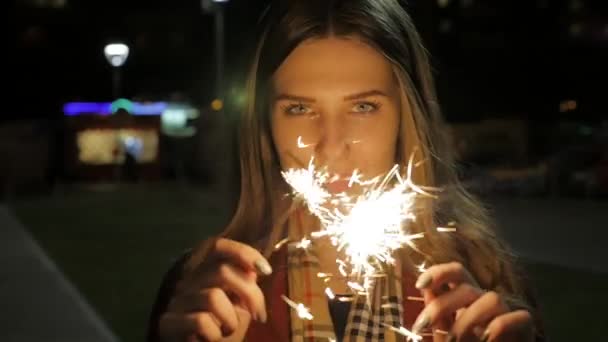 Hermosa joven sonriente sosteniendo un bengala. Escena nocturna. Joven chica sonriente sosteniendo chispa en su mano. Concepto de vacaciones — Vídeos de Stock